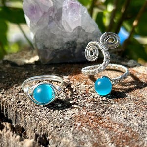 May include: Two silver wire wrapped rings with turquoise blue glass stones. The rings are on a wooden surface.