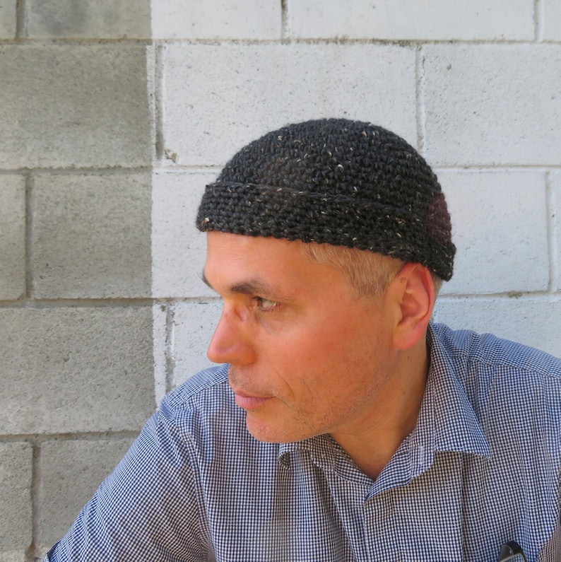 May include: A person wearing a black knit beanie with a flecked texture. The beanie has a simple design and is worn low on the forehead.