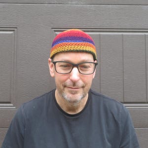 May include: A crochet beanie featuring horizontal stripes in vibrant colors including red, orange, yellow, green, blue, and purple. The hat has a rounded crown and a textured stitch pattern, constructed from yarn in a multi-colored rainbow design.