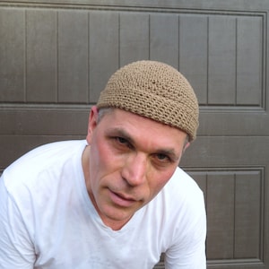 May include: A person wearing a tan crocheted beanie hat. The hat has a simple design and is made of a soft, textured yarn.