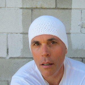 May include: A white crocheted beanie hat with a close-up of the person wearing it. The person has short brown hair and is wearing a white t-shirt.