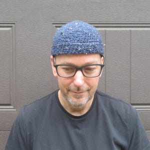 May include: A blue beanie hat with a speckled texture and a rolled brim. The hat is crafted from a thick, textured yarn featuring small flecks of white and tan throughout the blue base, creating a distinct, multi-tonal appearance on the fabric.