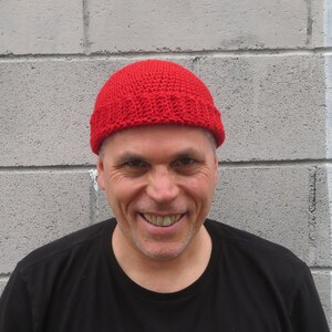 May include: A bright red, crocheted beanie hat. The hat is made of a textured yarn and has a folded brim. The hat is worn by a person wearing a black shirt.