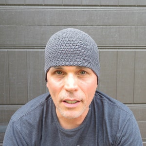 The model is wearing a slate gray, crocheted beanie that fits snugly and covers his ears.