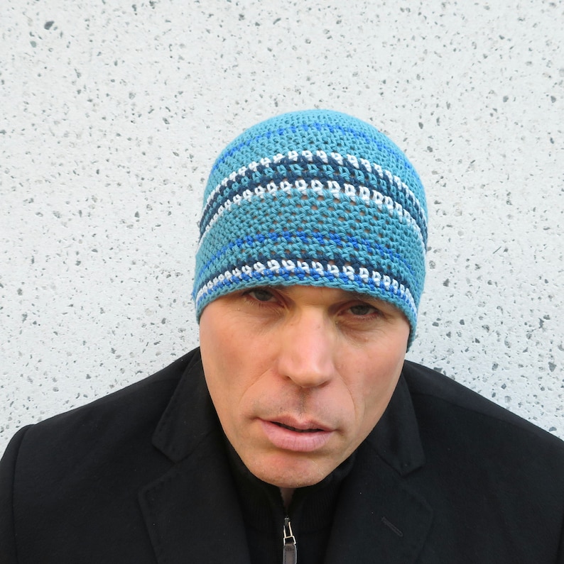 The model is wearing an ocean blue, crocheted beanie with stripes of light blue, dark blue and bright blue running at intervals throughout- it fits snugly and covers his ears.