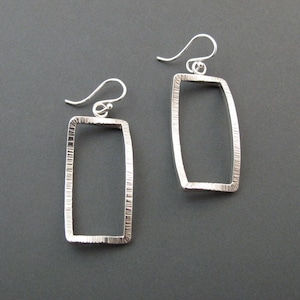May include: A pair of silver rectangular drop earrings with textured edges. The earrings have a simple, modern design with a hook closure. The background is a solid dark gray.