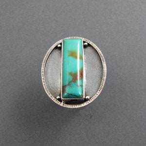 May include: A silver ring with a turquoise stone set in a rectangular frame. The ring is surrounded by a circular band with a textured edge.