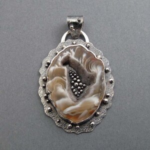 May include: A silver pendant with an oval, agate-like center in shades of brown, white, and gray. The pendant has a textured silver frame with small, round embellishments. A silver bail is at the top for a chain.