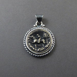 May include: A circular silver pendant featuring a central gold-toned relief of a person riding a horse jumping over a fence. The pendant is framed by a twisted silver rope border and includes a triangular silver bail at the top for attachment.