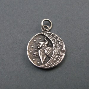 May include: A silver pendant featuring a detailed depiction of an angel with wings, standing within a crescent moon. The moon is decorated with a geometric pattern.