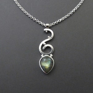 May include: A silver pendant necklace with a teardrop-shaped, iridescent gemstone. The pendant has a unique, swirling design with textured detailing and small, round accents. The chain is a simple, linked style.