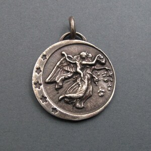 May include: Silver pendant featuring a detailed relief of an angel with wings outstretched, holding a small figure. The pendant is circular and has a star-shaped border.