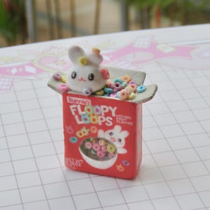 Op de afbeelding: Een miniatuur rode ontbijtgranendoos met het opschrift "Bunnie's Floopy Loops", een cartoonkonijn en een kom met kleurrijke ontbijtgranen. Een wit konijnfiguur met een bloemenkroon zit bovenop de doos, omringd door ontbijtgranen.