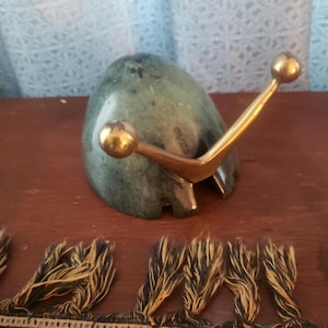 May include: A green stone letter holder with a brass accent. The letter holder has a rounded shape with a brass arm extending from the top, ending in two brass spheres. The base is a dark brown color.