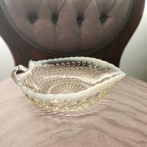 May include: A clear, leaf-shaped glass dish with a handle. The dish features a textured pattern of small circles around the rim and a sunburst design in the center. The glass has a slight opalescent quality.