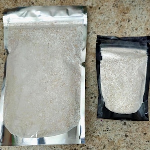 May include: Two stand-up pouches containing a light beige, granular substance. The larger pouch is silver with a clear front panel, while the smaller pouch features a black border with a clear front panel, both with resealable zipper tops.