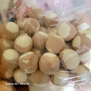 May include: A clear plastic bag filled with numerous round, light brown candies with a white, creamy coating on one side. The candies appear to be individually wrapped or coated, creating a textured appearance. The candies are likely a sweet treat.