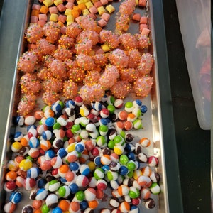 May include: A metal tray filled with various candies. The top section has small, square, pastel-colored candies. Below are pink, spherical candies covered in sprinkles. The bottom section is filled with colorful, round candies with a white stripe.