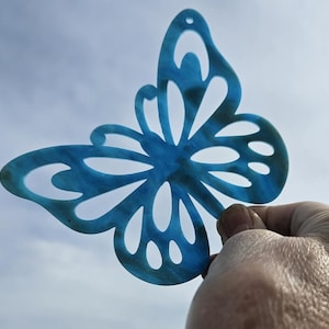 May include: A translucent blue butterfly-shaped ornament with cut-out details. The butterfly is held up against a cloudy sky. The ornament has a hole at the top for hanging.