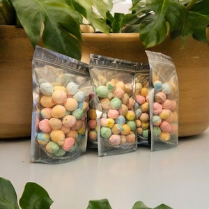 May include: Three clear plastic bags filled with pastel-colored candy coated chocolate eggs. The bags are sitting on a white surface.