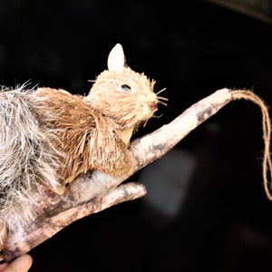 May include: A decorative squirrel ornament made of straw and faux fur, perched on a painted branch. The squirrel has a fluffy tail and is designed to hang. The ornament is a rustic, woodland-themed decoration.