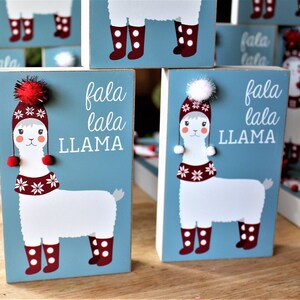 May include: Wooden blocks with a light blue background feature a white llama illustration wearing a red hat, scarf, and boots. The text "fala lala LLAMA" is printed in white. Each block has a red pom-pom on top.