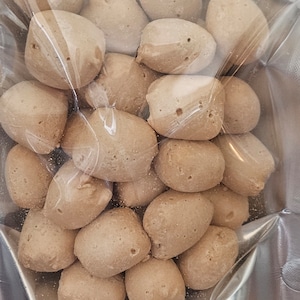 May include: A close-up of a bag of small, round, light brown food items. The food items are arranged in a random pattern and are visible through the clear plastic bag.