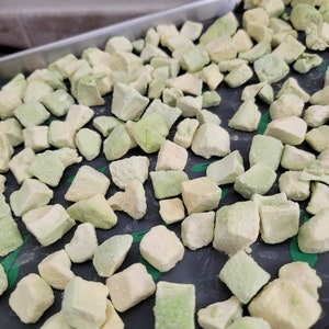 May include: A tray filled with numerous, irregularly shaped, light green and off-white cubes. The cubes are scattered across a dark surface, suggesting a food preparation or preservation process. The overall color palette is muted, with a focus on the natural tones of the food.