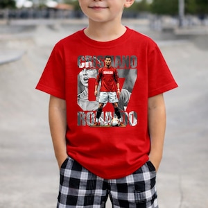 May include: A bright red short-sleeved t-shirt featuring a graphic print of soccer player Cristiano Ronaldo in a red jersey and white shorts. The design includes the text CRISTIANO RONALDO in bold lettering over a black and white background.