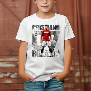 May include: A white short-sleeved t-shirt featuring a graphic print of Cristiano Ronaldo in a red jersey and white shorts. The design includes the text CRISTIANO RONALDO in bold black letters, a black and white portrait, and a soccer ball at the feet.