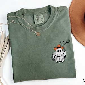 K&ouml;nnte beinhalten: Moosgr&uuml;nes T-Shirt mit einem Cartoon-Geist, der einen Cowboyhut tr&auml;gt und ein Lasso h&auml;lt. Eine goldene Halskette und ein brauner Hut sind ebenfalls im Bild zu sehen. Das T-Shirt hat ein Etikett mit der Aufschrift "Comfort Colors".