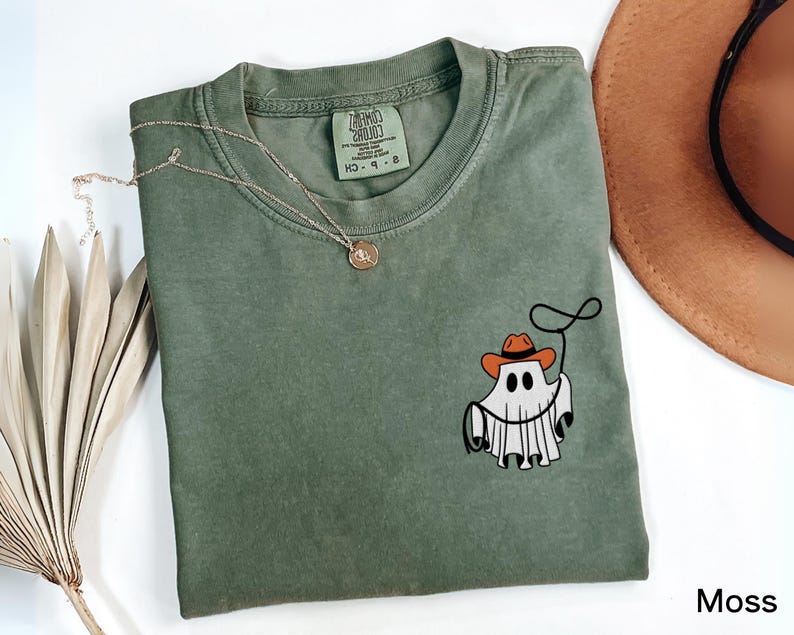 K&ouml;nnte beinhalten: Moosgr&uuml;nes T-Shirt mit einem Cartoon-Geist, der einen Cowboyhut tr&auml;gt und ein Lasso h&auml;lt. Eine goldene Halskette und ein brauner Hut sind ebenfalls im Bild zu sehen. Das T-Shirt hat ein Etikett mit der Aufschrift "Comfort Colors".