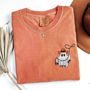 K&ouml;nnte beinhalten: Korallfarbenes T-Shirt mit Rundhalsausschnitt und einer kleinen Grafik eines Geistes mit Cowboyhut und Lasso. Eine goldene Halskette und ein brauner Cowboyhut sind ebenfalls sichtbar.