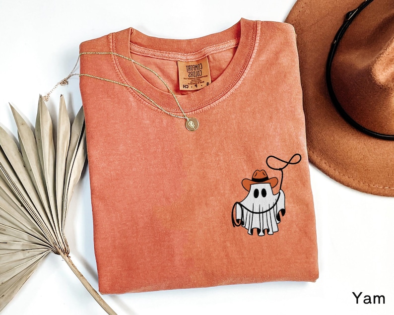 K&ouml;nnte beinhalten: Korallfarbenes T-Shirt mit Rundhalsausschnitt und einer kleinen Grafik eines Geistes mit Cowboyhut und Lasso. Eine goldene Halskette und ein brauner Cowboyhut sind ebenfalls sichtbar.