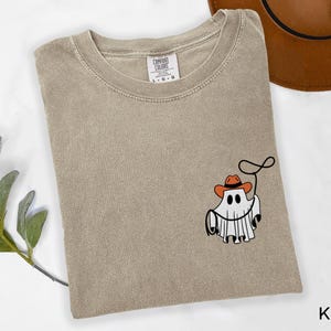 K&ouml;nnte beinhalten: Khaki-farbenes T-Shirt mit einer Grafik eines Geistes, der einen Cowboyhut tr&auml;gt und ein Lasso h&auml;lt. Das Shirt hat einen Rundhalsausschnitt und kurze &Auml;rmel. Das Markenetikett ist auf der oberen R&uuml;ckseite des Shirts sichtbar. Das Wort "Khaki" ist unten rechts aufgedruckt.