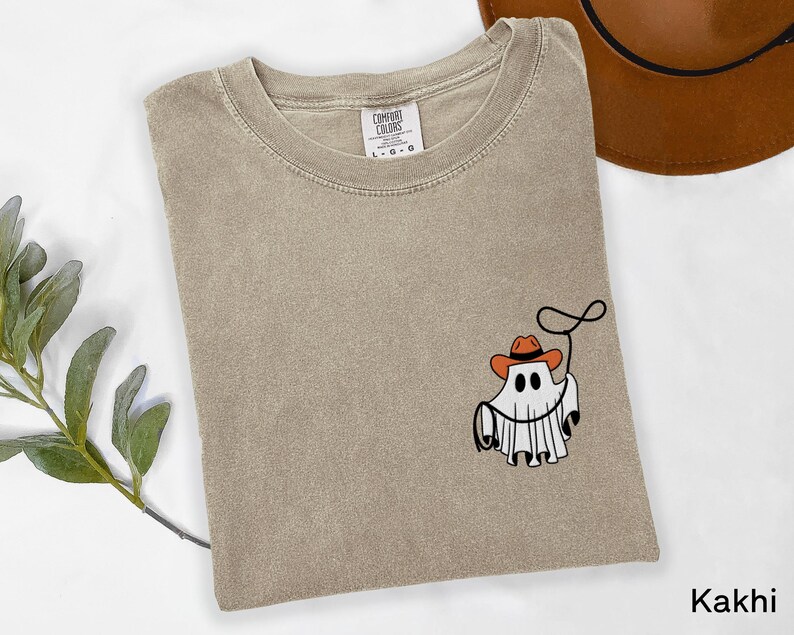 K&ouml;nnte beinhalten: Khaki-farbenes T-Shirt mit einer Grafik eines Geistes, der einen Cowboyhut tr&auml;gt und ein Lasso h&auml;lt. Das Shirt hat einen Rundhalsausschnitt und kurze &Auml;rmel. Das Markenetikett ist auf der oberen R&uuml;ckseite des Shirts sichtbar. Das Wort "Khaki" ist unten rechts aufgedruckt.