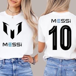 May include: White t-shirt with a black graphic of the letter "M" and the word "MESSI" in blue and black. The back of the shirt has the number "10" in black with the word "MESSI" in blue and black.