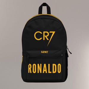 May include: Black backpack with yellow accents and text. The front features the text "CR7" above "GOAT" and "RONALDO" in yellow. The backpack has a top handle, a front pocket, and a side mesh pocket.