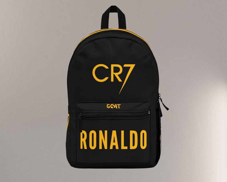 May include: Black backpack with yellow accents and text. The front features the text "CR7" above "GOAT" and "RONALDO" in yellow. The backpack has a top handle, a front pocket, and a side mesh pocket.