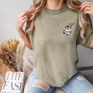 K&ouml;nnte beinhalten: Olivgr&uuml;nes T-Shirt mit einem Cartoon-Geist, der einen Cowboyhut tr&auml;gt und ein Lasso h&auml;lt. Die Grafik befindet sich auf der linken Brust. Das Shirt wird mit hellblauen Jeans kombiniert. Ein Buch und eine Glasflasche befinden sich im Hintergrund.