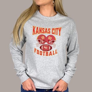 May include: Gray long-sleeve t-shirt with a red, yellow, and white graphic of two football helmets and a football with the text "KANSAS CITY 19 60 FOOTBALL".