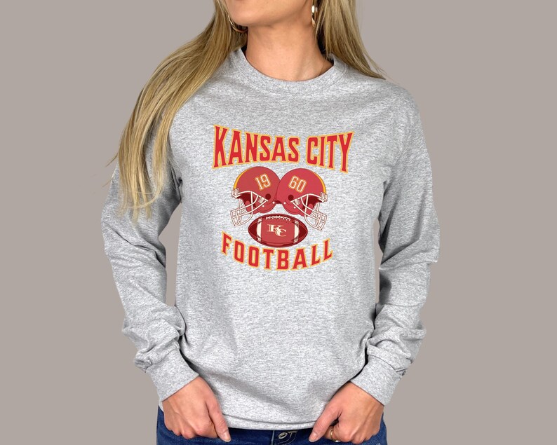 May include: Gray long-sleeve t-shirt with a red, yellow, and white graphic of two football helmets and a football with the text "KANSAS CITY 19 60 FOOTBALL".