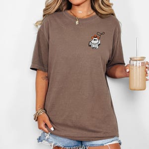 K&ouml;nnte beinhalten: Braunes T-Shirt mit einer kleinen Grafik eines Cartoon-Geistes, der einen Cowboyhut tr&auml;gt und ein Lasso h&auml;lt. Das Shirt ist einfarbig und hat eine lockere Passform. Das Model tr&auml;gt Jeansshorts und h&auml;lt ein Glas mit einem Strohhalm.