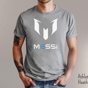 May include: A gray t-shirt with a white graphic of the letter "M" and the word "MESSI" in blue and white letters.