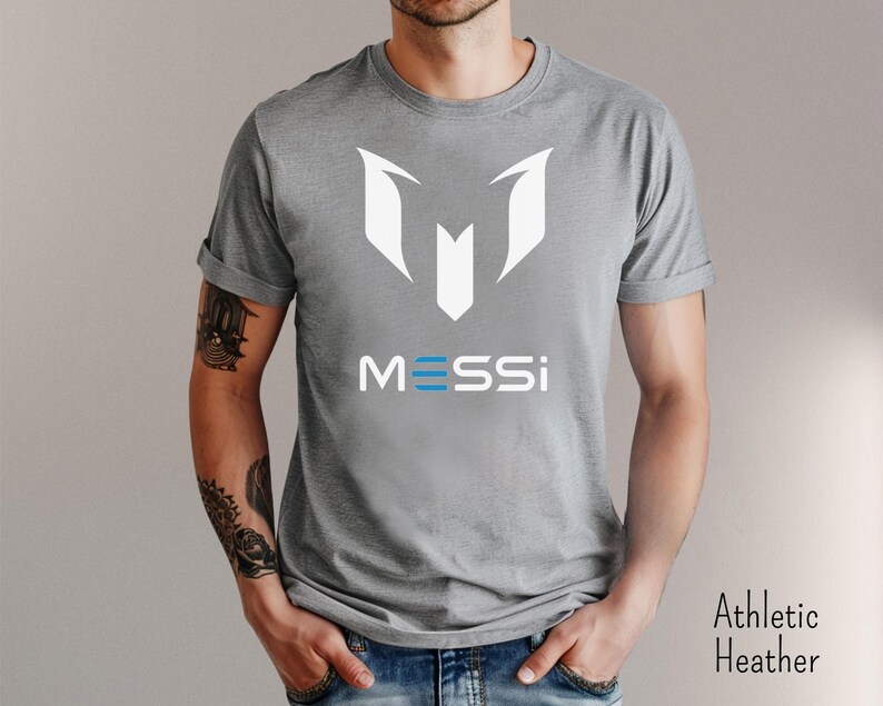 May include: A gray t-shirt with a white graphic of the letter "M" and the word "MESSI" in blue and white letters.