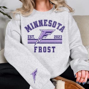 Pode incluir: Uma camisa de moletom cinza com um gr&aacute;fico roxo de um raio e o texto "Minnesota Est. 2023 Frost".