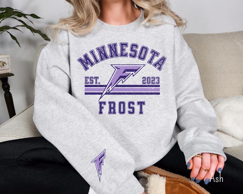 Pode incluir: Uma camisa de moletom cinza com um gr&aacute;fico roxo de um raio e o texto "Minnesota Est. 2023 Frost".