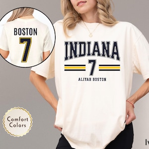 May include: Ivory-colored t-shirt featuring the word "INDIANA" in navy blue block letters with a white outline, and the number "7" in navy blue with a yellow outline. The name "ALIYAH BOSTON" is printed below. A smaller image shows the back of the shirt with "BOSTON" and "7".