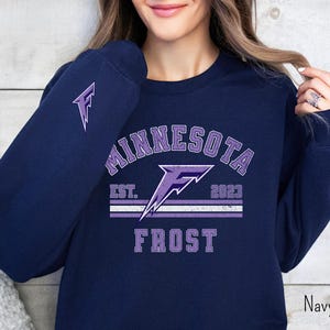 Pode incluir: Camisola azul marinho com um design gr&aacute;fico roxo que diz "Minnesota Frost Est. 2023" com um raio roxo.