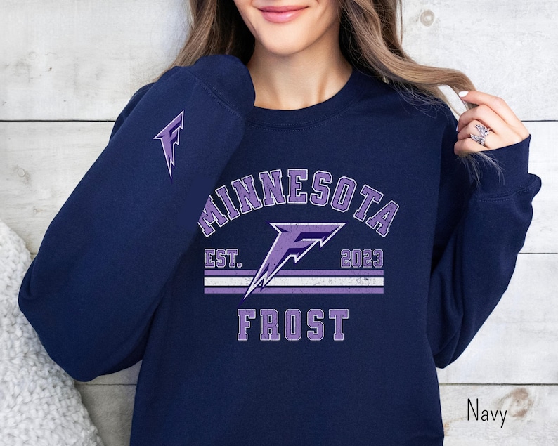 Pode incluir: Camisola azul marinho com um design gr&aacute;fico roxo que diz "Minnesota Frost Est. 2023" com um raio roxo.